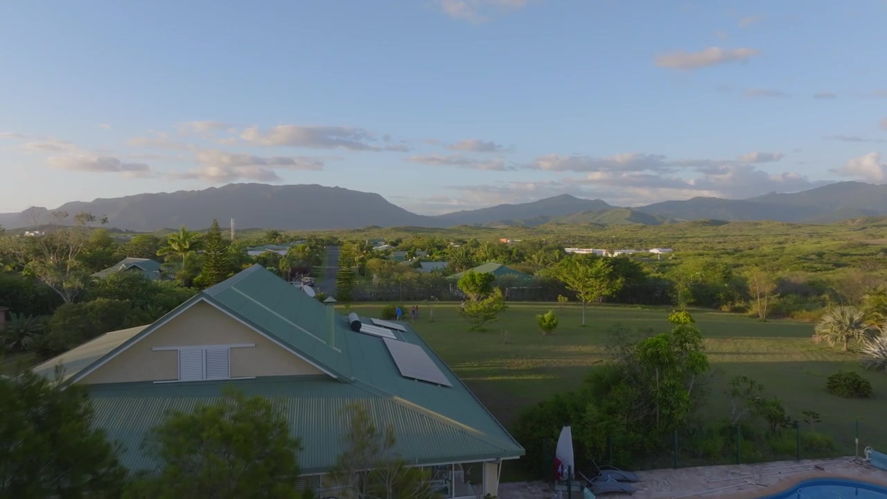 Lavtflyvende drone stiger op over et landbrugshegn for at afsløre en tropisk landsby, fjerne bjerge og en stor have, hvor folk samles til en udendørs aktivitet