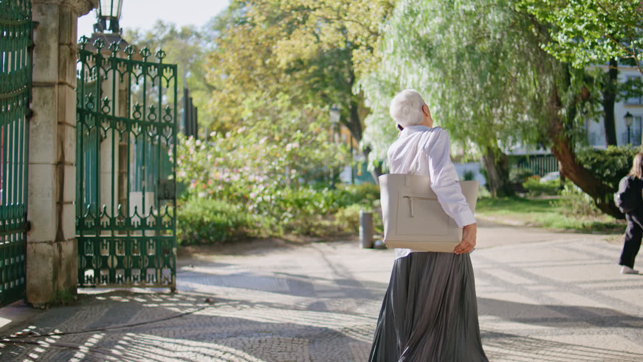 Urban pensioner speaking cellphone in sunny park. Stylish grey hair lady calling