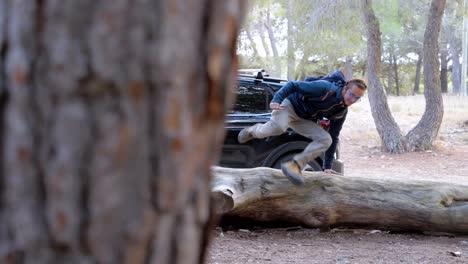 tracking shot of a man picking up his bag and jumping over a fallen tree ready for a hike