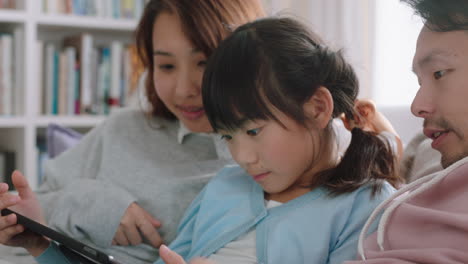 happy asian family using tablet computer little girl watching entertainment with parents playing game on touchscreen device browsing online relaxing on sofa together at home 4k footage