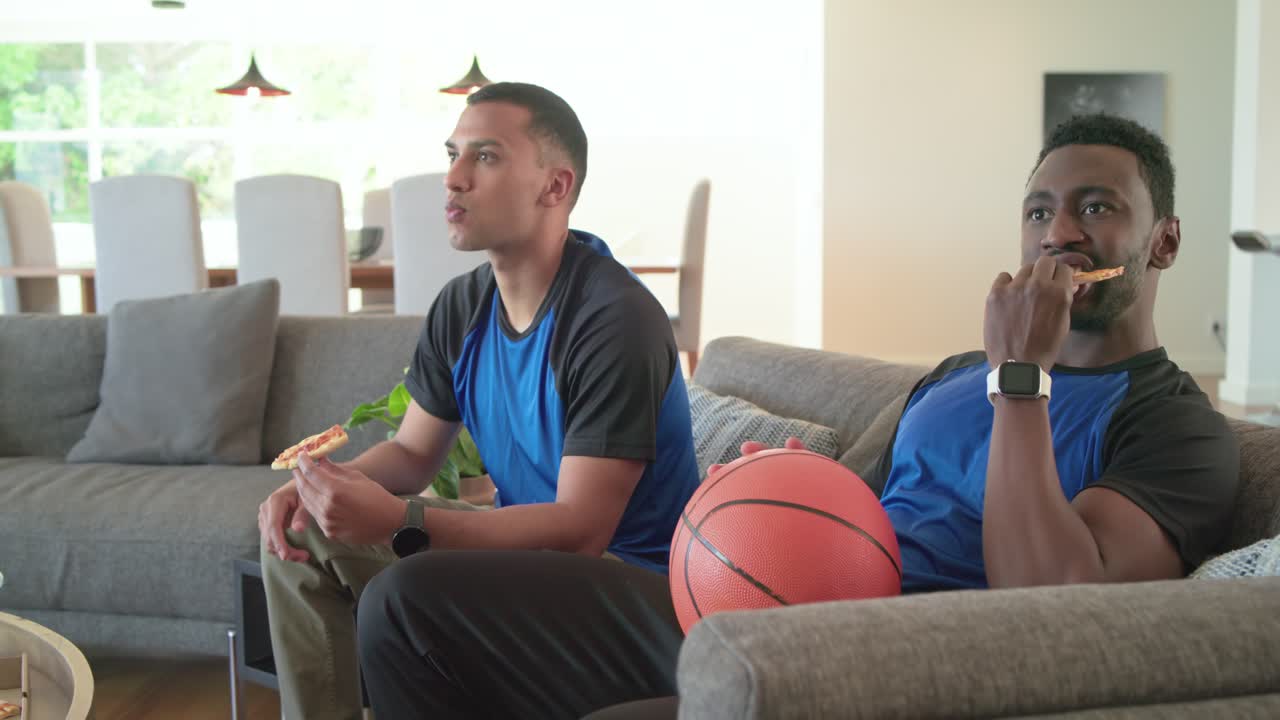 En gruppe mannlige venner ser på sportssending, roterer en basketball og spiser pizza hjemme.