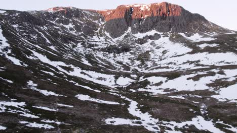 Aerial-drone-footage-slowly-rising-to-reveal-a-steep,-dramatic-and-imposing-mountain-cliff-face-covered-snow-in-winter-at-sunrise-with-boulders-and-rocks