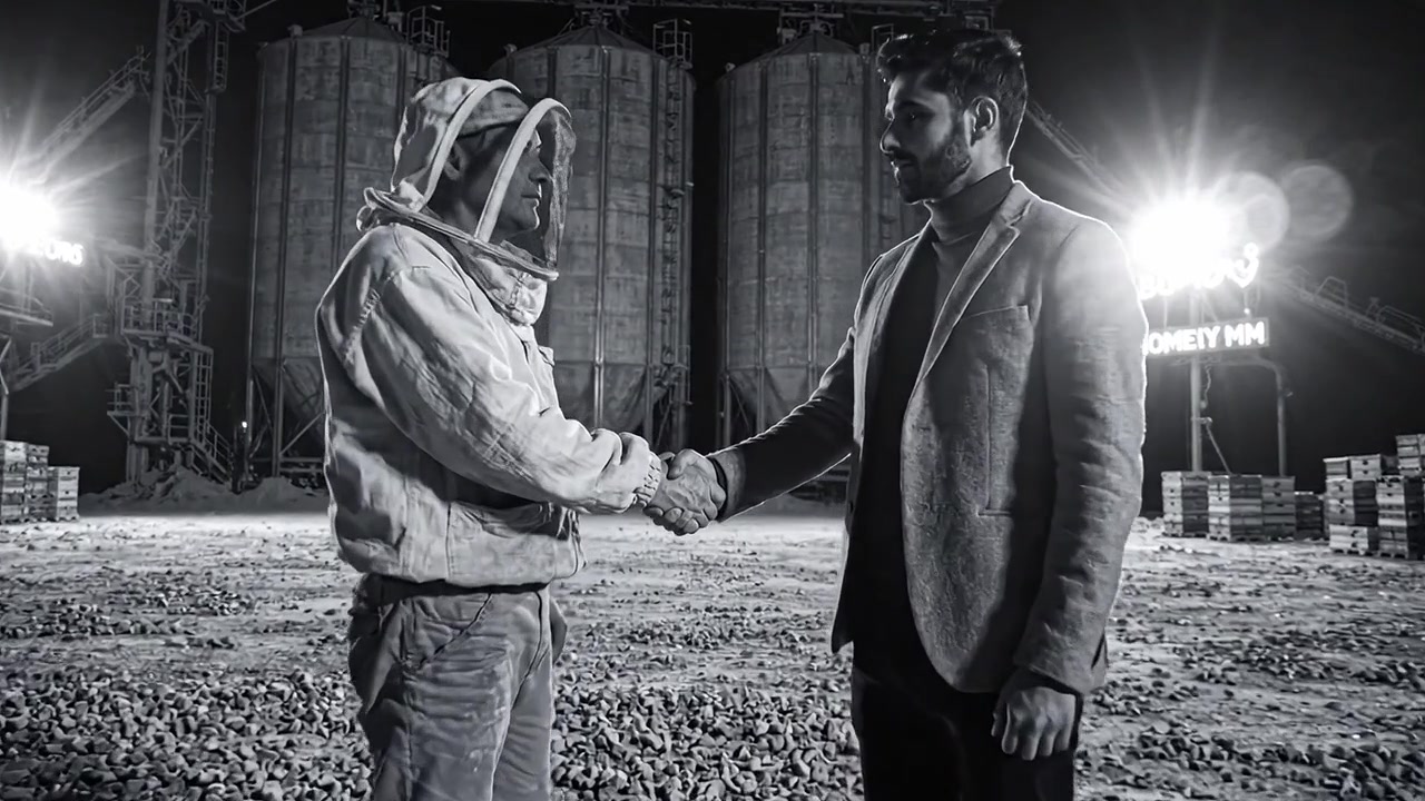 Ett avgörande handslag mellan två individer framför industrisilos, som symboliserar samarbete och överenskommelse i en skarp svartvit miljö