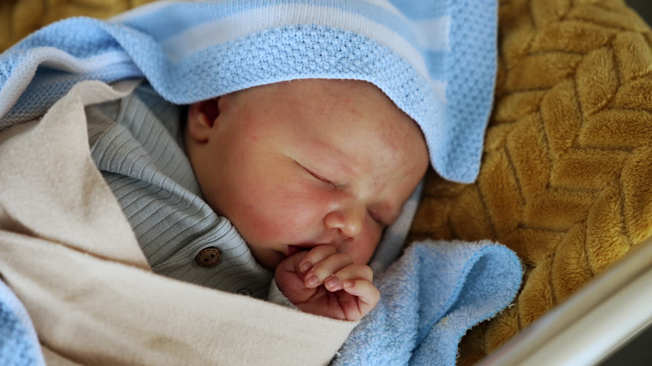 Newborn child sleeping in a crib holding his hand fold in fist at the mouth. Infant after delivery in a hospital ward. Close up.