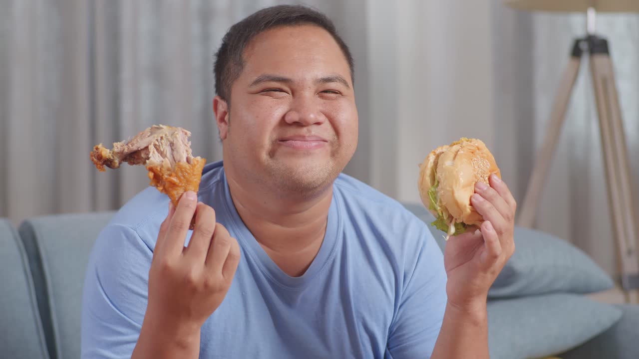 한 아시아 남자가 김과 햄버거를 들고 집에서 거실 소파에서 패스트푸드를 먹는 동안 카메라에 미소 짓고 있습니다.