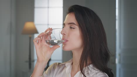 cinematic shot of young smiling woman is drinking fresh pure water from a transparent glass in living room at home. she maintains her body healthy and feels well being.