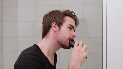 un joven adulto recortándose y afeitándose la barba frente al espejo en el baño.