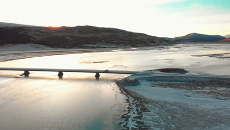 Vehicle-travelling-across-Lagarfljót-bridge,-East-Iceland-crossing-sparkling-sunset-river-aerial-orbiting-view