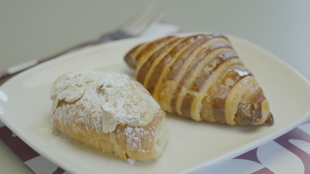 Close-up of two pieces of fresh pastry on a white plate: one glazed croissant and another covered with icing sugar
