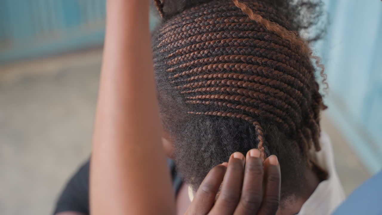 Nattes collées terminées mettant en valeur des rangées nettes sur le cuir chevelu, mains ajustant les bords et fixant les extrémités, coiffure protectrice élégante pour un usage quotidien, posture confiante de la cliente, ambiance de salon de marché en plein air, touche finale d'un styliste qualifié