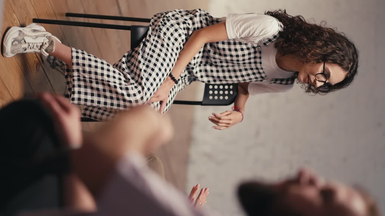 Vertical video: A brunette girl with curly hair wearing glasses in a checkered jumpsuit sits on a chair and talks about her problem in group therapy. Expressing your thoughts and problems in group therapy