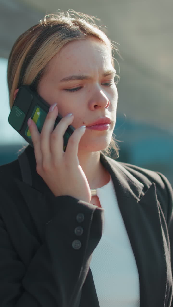 Lady pauses call, lowers phone to chest, takes deep breath while standing in urban setting with blurred city buildings and cars in background, dressed in professional attire