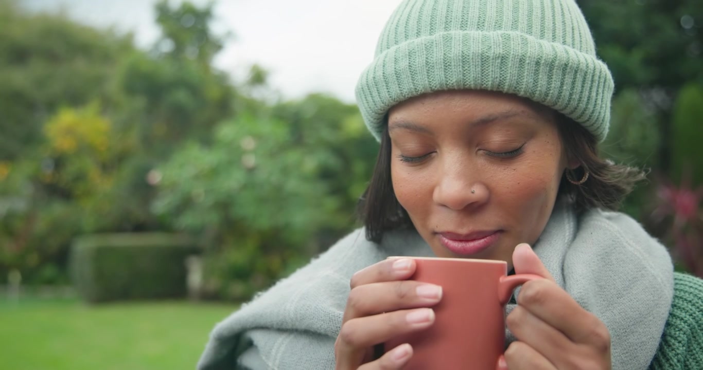 Donna afroamericana con un berretto, seduta in un cortile fresco, che tiene una tazza, inala l'aroma e sorseggia una bevanda calda