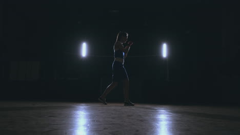 a beautiful woman boxer trains in a dark gym and works out punches in slow motion. side view. steadicam shot