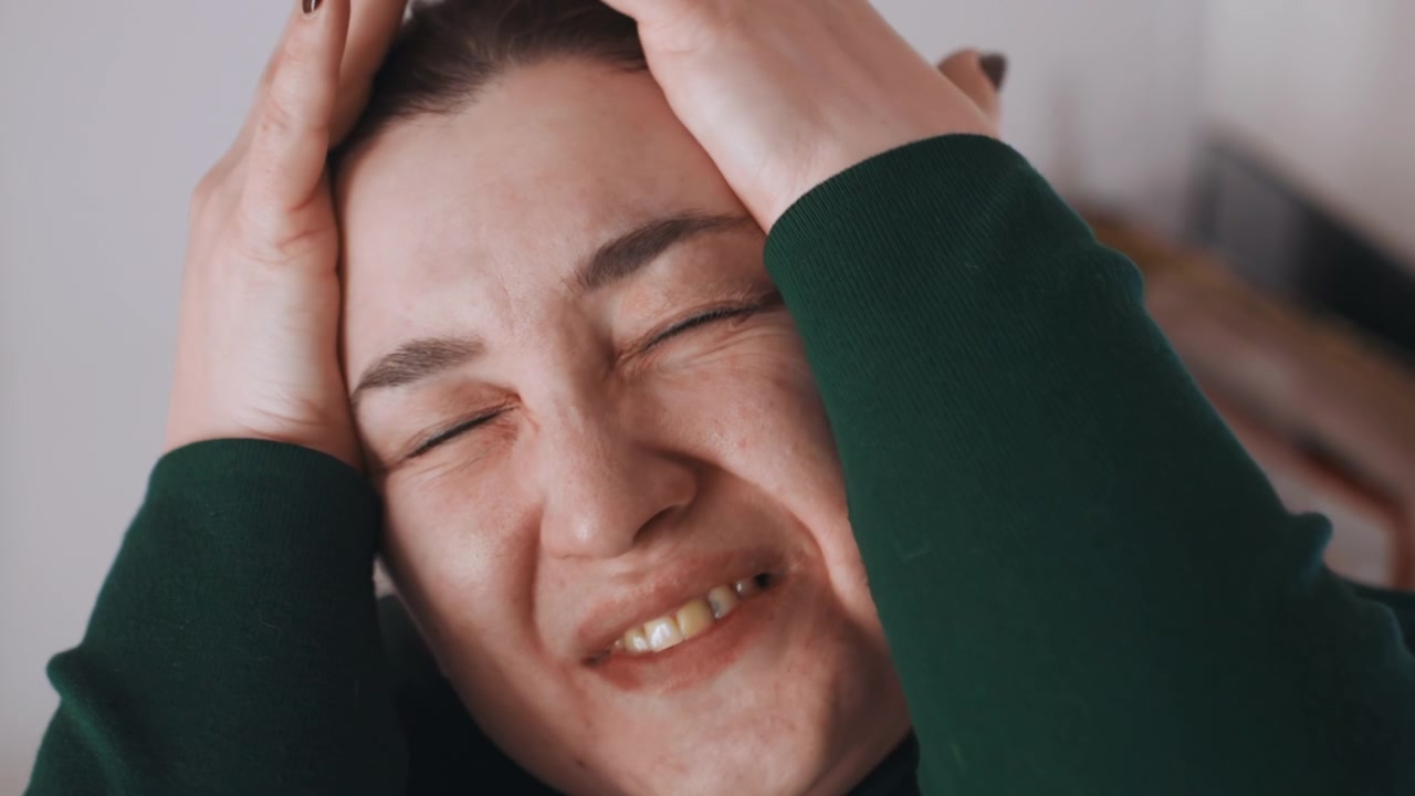 Donna bianca con intenso dolore facciale e smorfia, un primissimo piano cattura occhi lacrimosi, mascella serrata, mani che premono le tempie, sofferenza acuta e sensibilità alla luce, con accenni di un interno domestico sullo sfondo.
