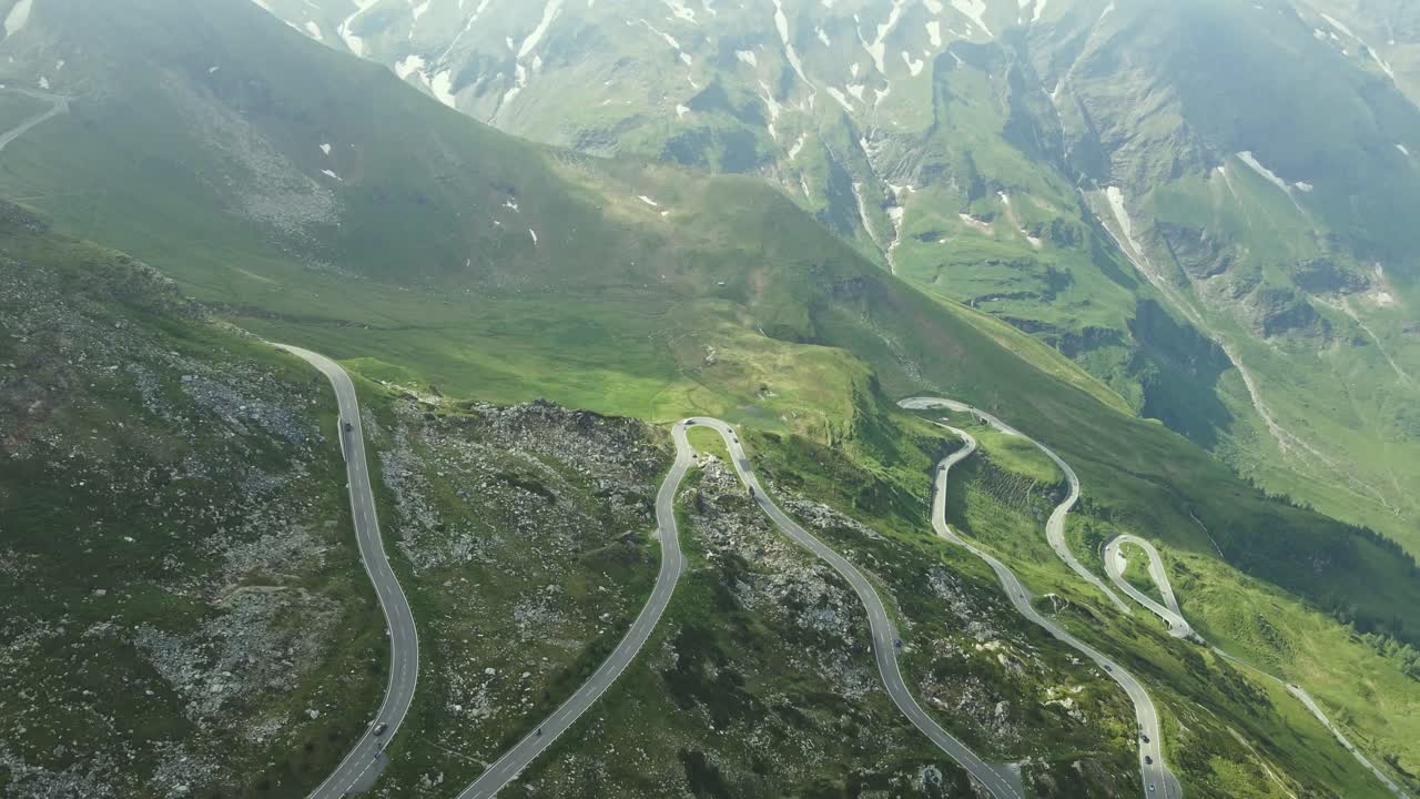 Aerial view of a winding mountain road snaking through a lush alpine landscape. The dramatic switchbacks and rugged terrain highlight the elevation and natural beauty of the high-altitude environment