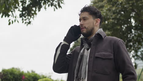 caucasian male model talking at phone having a business call outdoors with tree in background during winter season wearing gloves and scarf modern clothing outfits