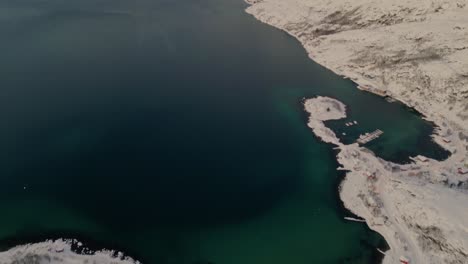 Panoramic-aerial-view-from-a-snow-covered-coastal-village-to-the-beautiful-emerald-waters-of-the-fjords