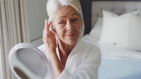 Happy-senior-caucasian-woman-sitting-on-bed-in-bedroom,-looking-at-mirror,-touching-her-hair