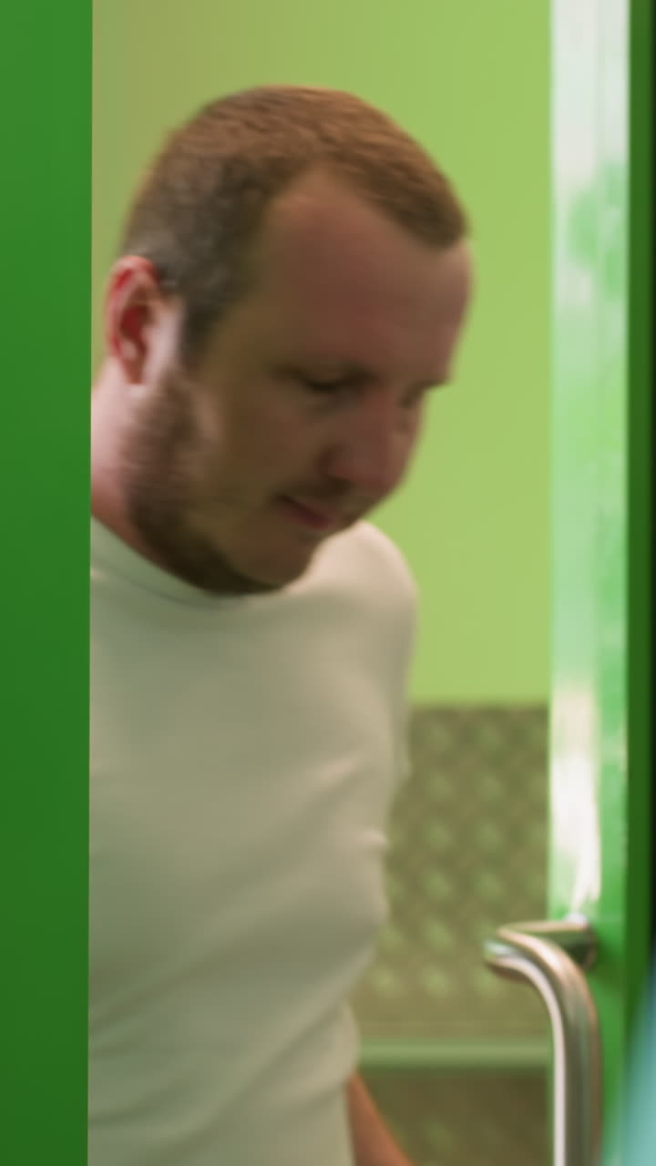 A close view of a man coming out through a green glass door with green frame on white polo, with a white object placed before him