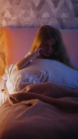 blonde little girl hugging large pillow wipes away tears sitting alone on bed by toy tent with glowing night lamp inside in bedroom at night slow motion