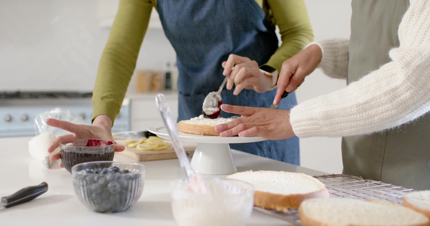 Duas mulheres empilhando, pegando compota com colher e espalhando sobre um bolo em um suporte, decorando em uma cozinha doméstica