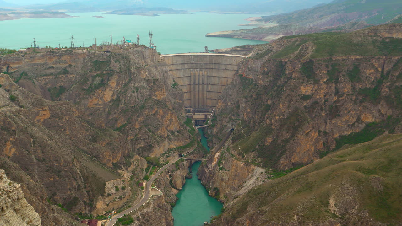 Dik dağ uçurumları arasında devasa bir beton barajın, turkuaz nehrin, hidroelektrik santralinin ve su yönetimi altyapısının geniş açılı görüntüsü; kanyonun içinden turkuaz renkli nehir akıyor