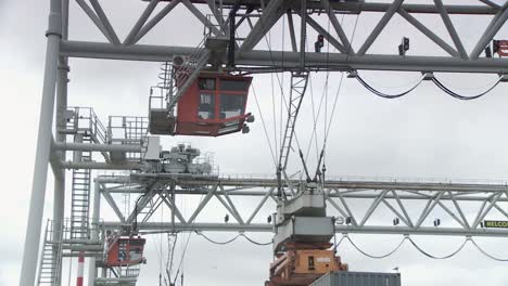 A-crane-at-a-harbor-with-a-red-cabin-with-an-individual-inside,-cables-attaching-the-cabin-to-the-crane-on-a-metal-structure-with-stairs-and-railings-against-a-cloudy-sky-backdrop