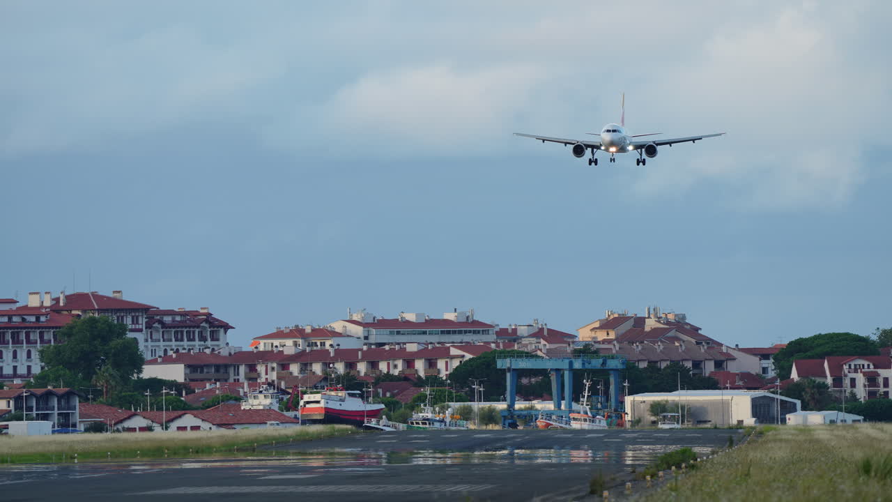 Commercial passenger jet approaching airport runway with town buildings and boats in background