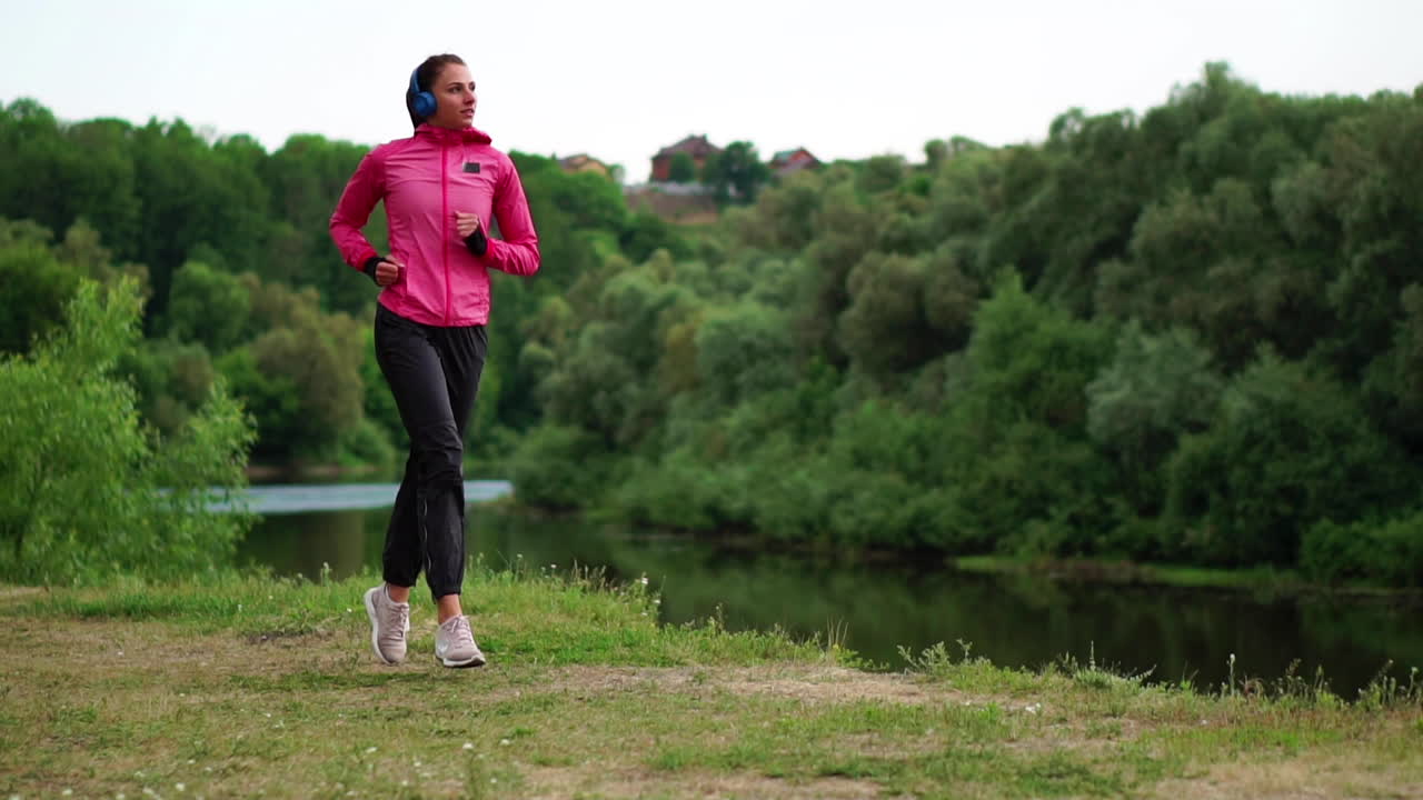 A girl in a pink jacket and black pants runs near the river in headphones preparing for the marathon