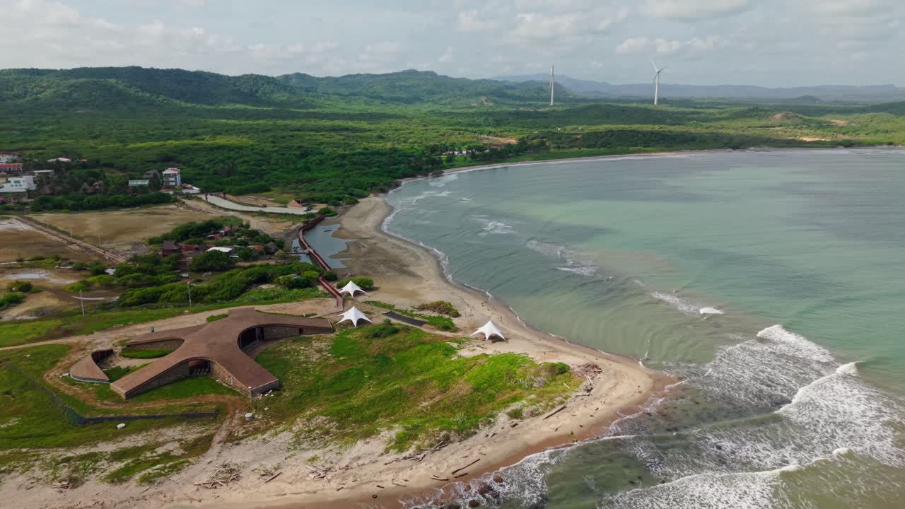 Slow motion aerial view of waves crashing on a tropical beach with lush vegetation and modern architecture, colombia