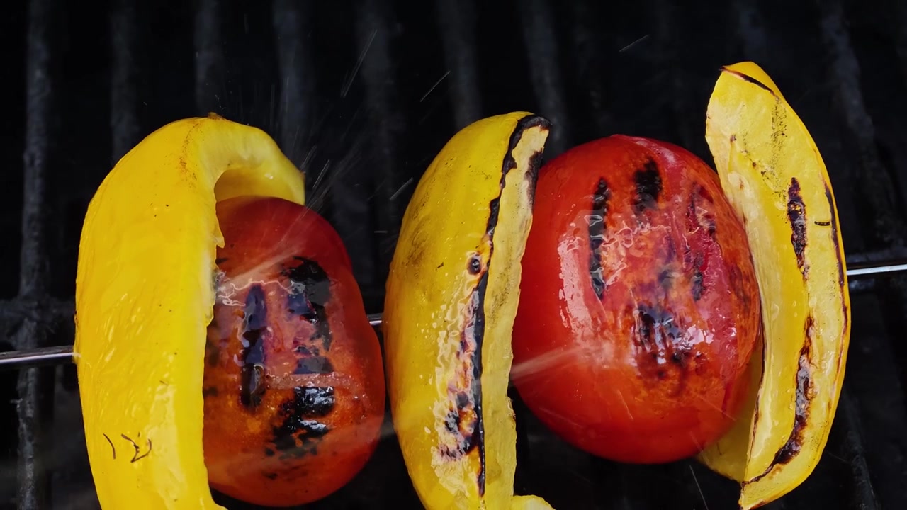 Primo piano al rallentatore di spiedino di verdure grigliate con pomodori rossi interi, fette di peperone giallo e olio d'oliva che schizza su una griglia BBQ nera, riprese macro cinematografiche dall'alto di cibo in 4K al tramonto!