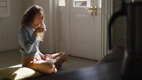 Caucasian-woman-sitting-on-floor-drinking-cup-of-coffee-using-laptop-in-sunny-cottage-living-room