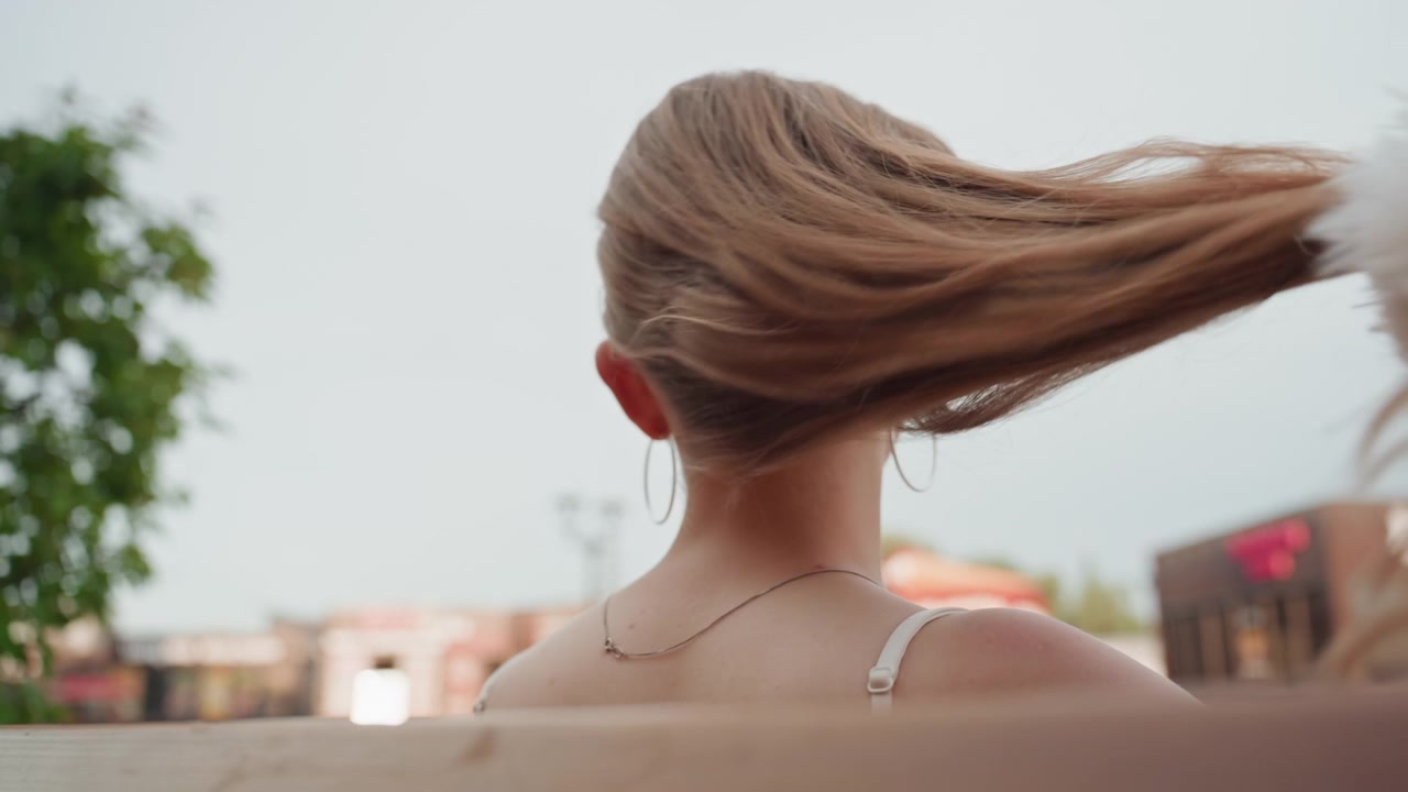 Donna con lunghi capelli legati a coda di cavallo siede su una panchina all'aperto, sistemando il suo elastico per capelli bianco piumato e lasciando cadere i capelli sciolti. Lo sfondo mostra edifici sfocati e vegetazione