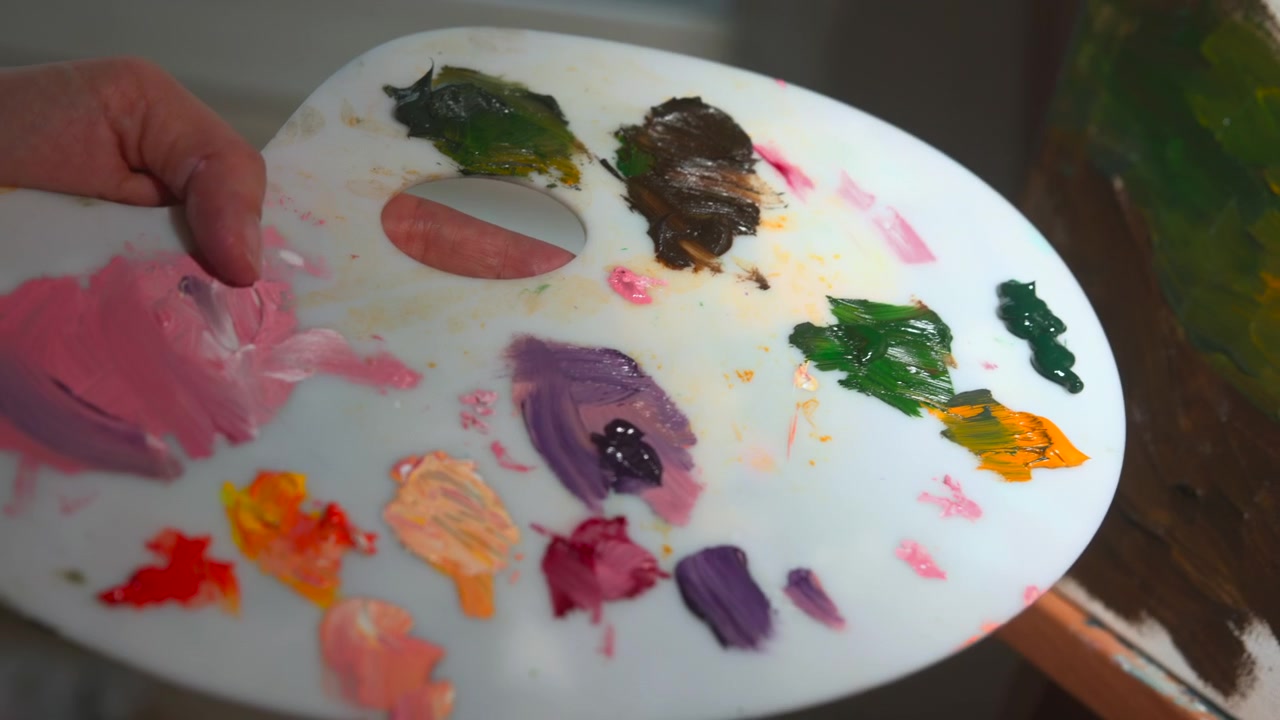 Una vista ravvicinata mostra una mano che usa un pennello per applicare pittura densa verde, marrone e oro su una tela bianca con una luce interna soffusa