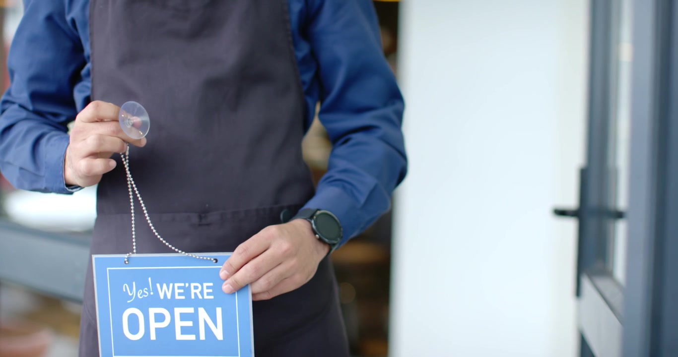 African man in apron mounting blue OPEN sign using suction cup at storefront, preparing for opening
