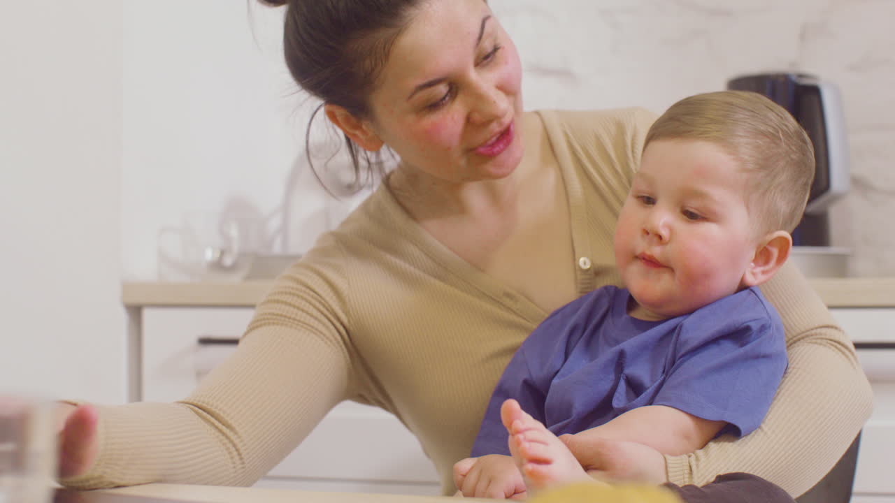 Young Mother Holding And Kissing Her Baby Boy While Working With Laptop Computer At Home
