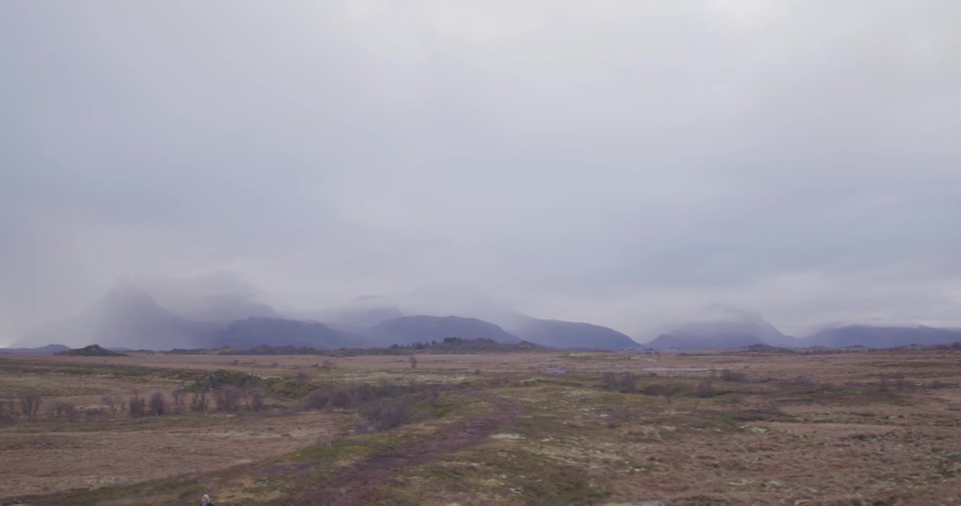 Desolate marshy field with leafless shrubs, rugged terrain, and distant mountains partially obscured by low-hanging clouds under an overcast sky, creating a somber, moody scene.