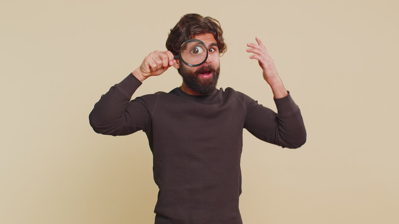 Portrait of young male investigator holding magnifying glass in amazement against beige background