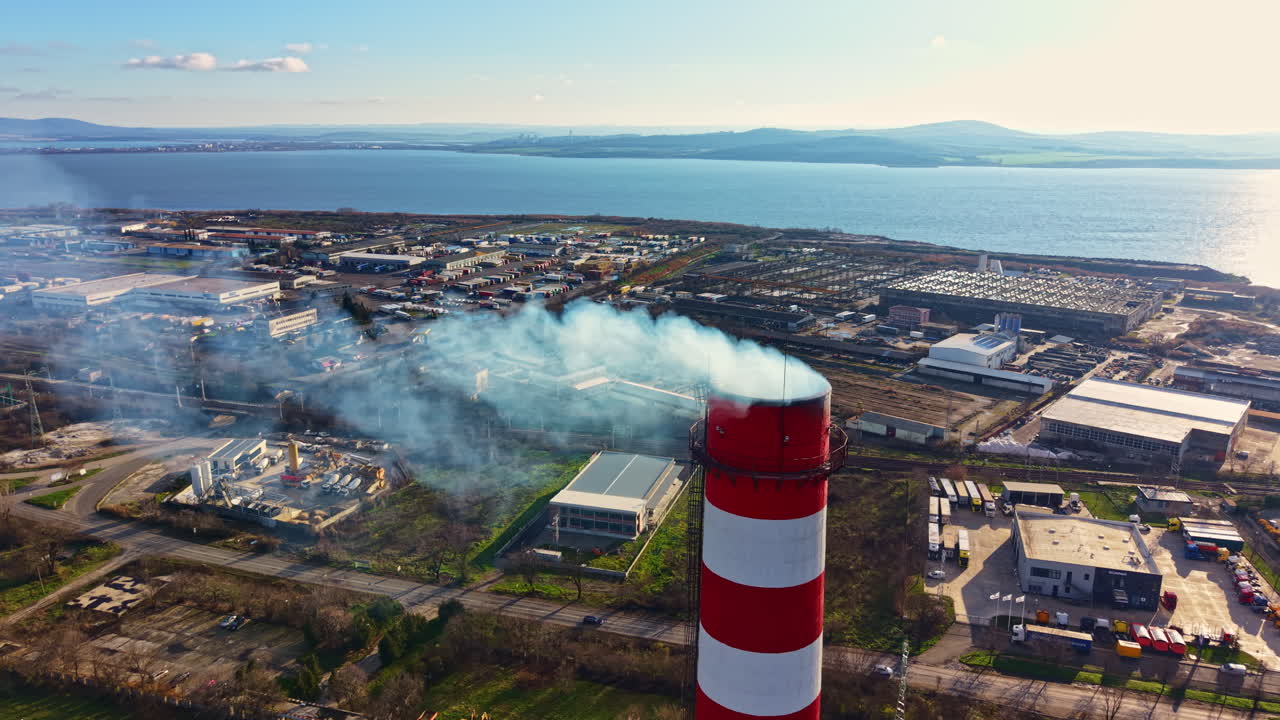 Del fumo si alza da un’alta ciminiera in una zona industriale costiera durante il giorno, con fabbriche e acqua sullo sfondo