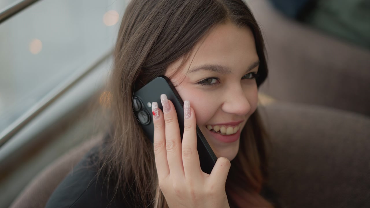 Giovane donna seduta su un divano in tessuto neutro accanto a una finestra luminosa, che sorride mentre parla al telefono con postura rilassata, leggera inclinazione della testa, sguardo concentrato e un'atmosfera domestica calma