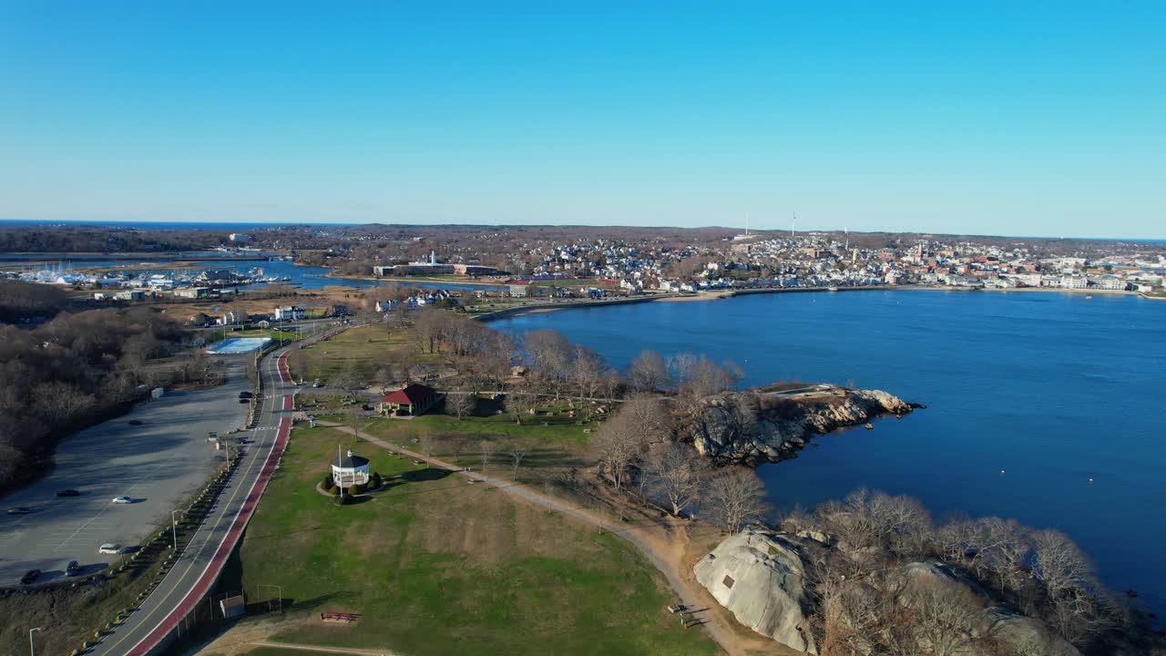 Letecký pohled na pobřeží Gloucesteru, Massachusetts s cyklostezkou, baseballovým hřištěm, skalnatým břehem a klidnou modrou vodou. Malebný pobřežní park a přímořské město pod jasnou oblohou. Přibližující se záběr.