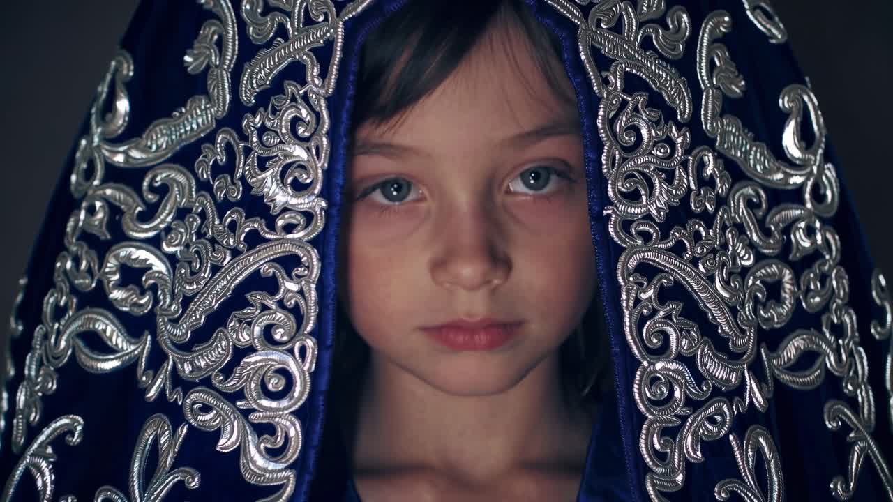 Young girl with blue eyes, adorned in a richly embroidered blue cloak, gazes intently, revealing emotions through intricate fabric details and soft lighting, creating a captivating visual narrative
