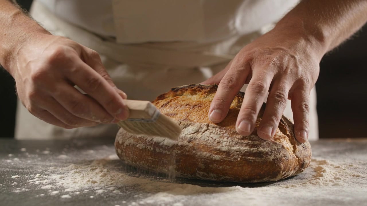 Placing hands on artisan loaf, baker brushing flour off crust with brush at bakery, white coat