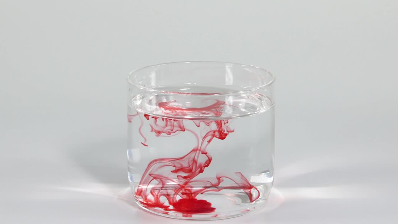 Red dye creates intricate patterns as it disperses in a clear glass of water.