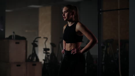 Young-woman-resting-after-her-workout-and-looking-down.-Female-athlete-taking-rest-after-fitness-training-in-dark-gym