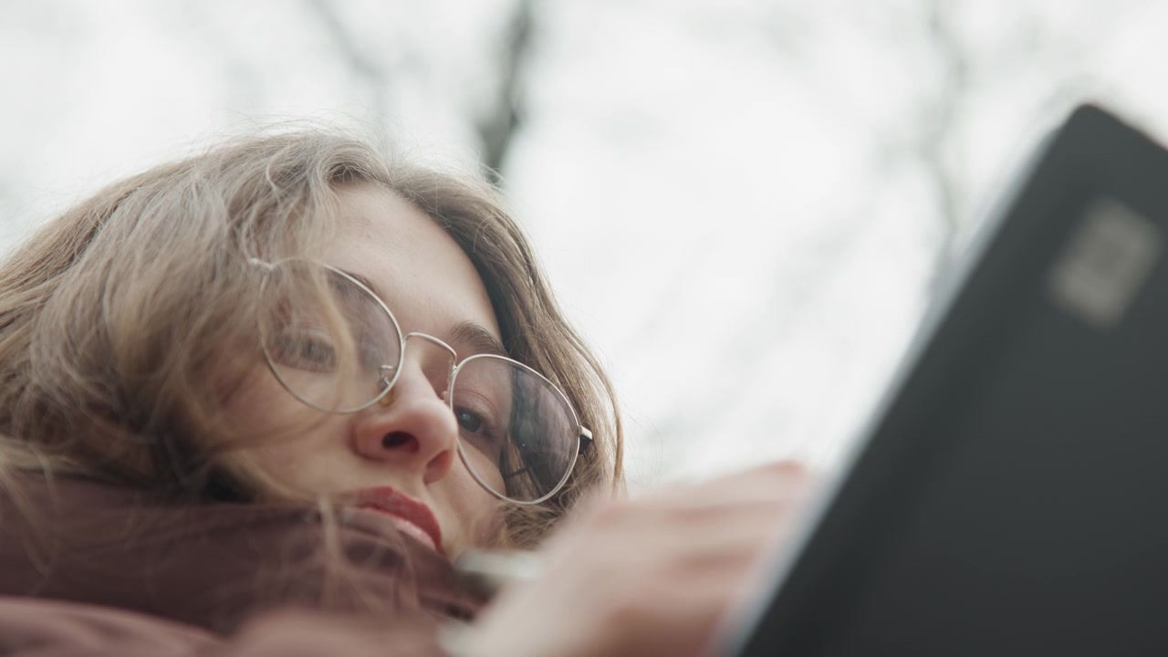 Young white woman scrolling phone outdoors, brown coat and round glasses, overcast sky and bare trees background, closeup lowangle shot, introspective expression, student researching messages