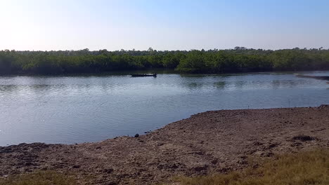 Image-of-a-river-at-low-tide-with-a-fisherman-passing-by-in-a-canoe,-a-river-bank-with-dry-mud-on-the-other-side-the-green-of-the-extensive-mangroves