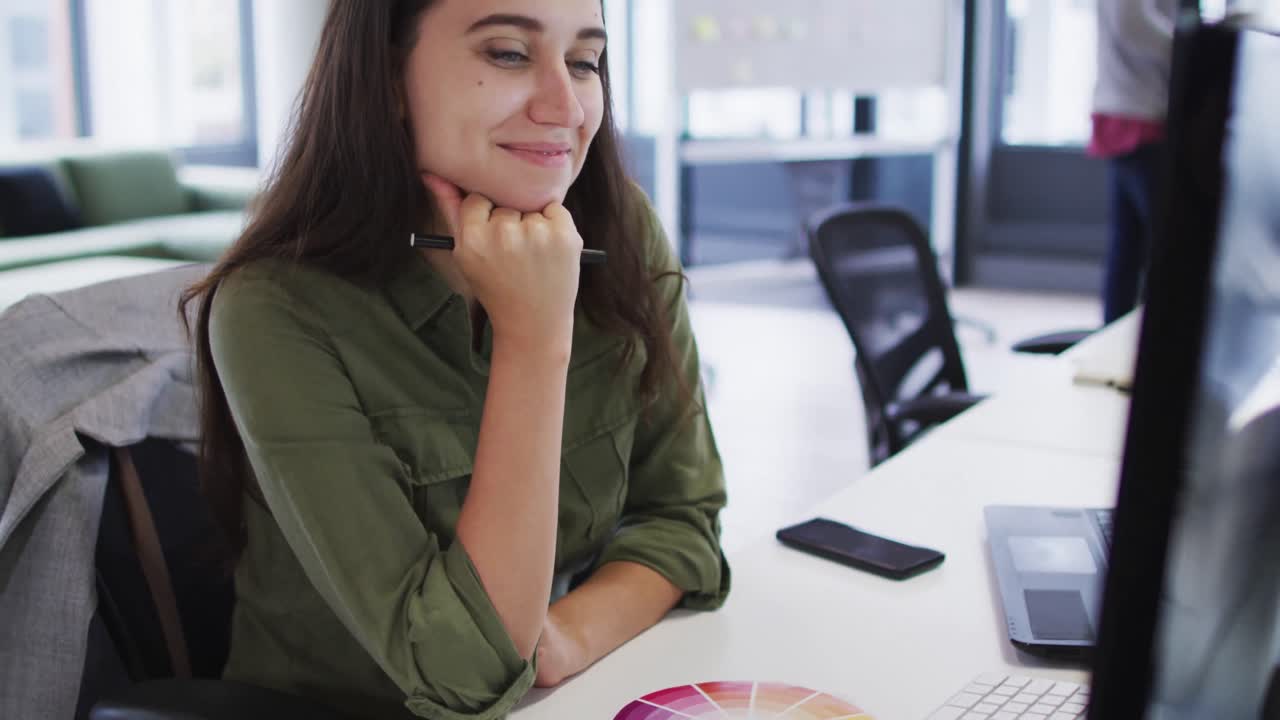 mujer de negocios caucásica sentada en un escritorio mirando el monitor de la computadora y sonriendo