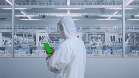 back view of asian male scientist use mock up green screen smartphone while standing in modern lab factory with automated robots